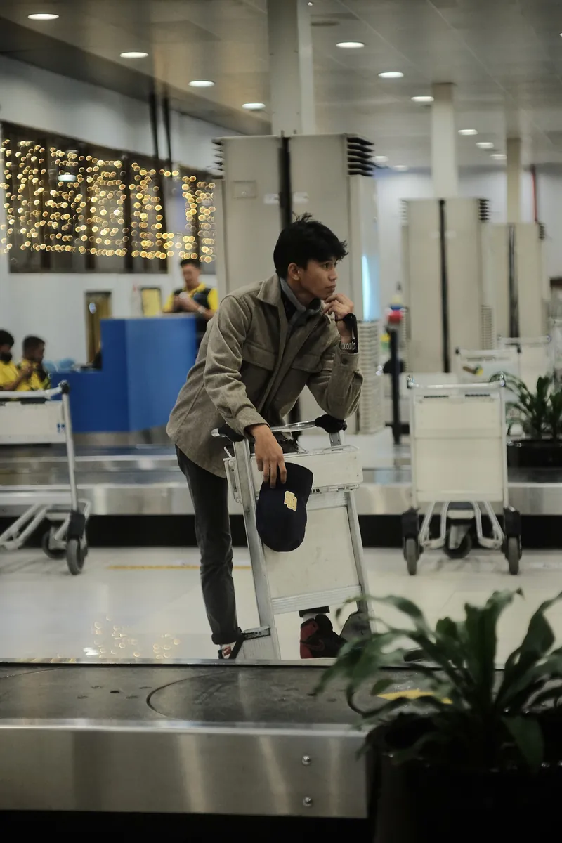 Homem de casaco em pé, apoiado em um carrinho de bagagem ao lado da esteira de um aeroporto.
