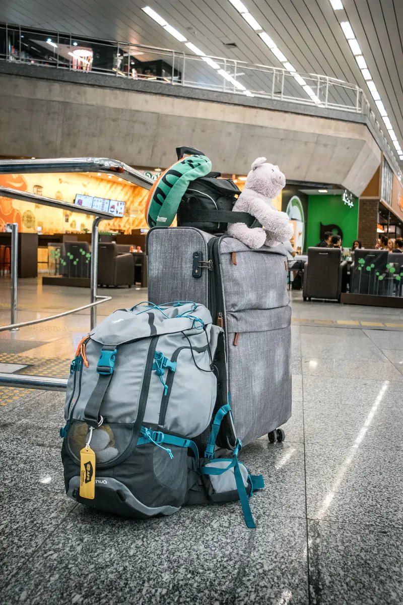 Mala cinza com uma mochila e um ursinho de pelúcia em cima, no chão de um saguão de aeroporto.