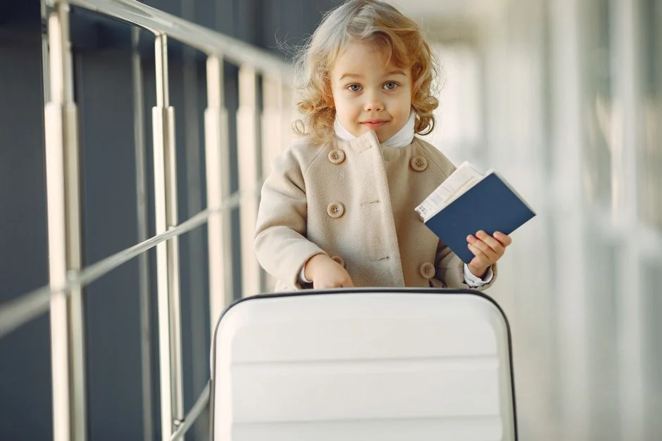 Criança pequena segurando um passaporte fechado ao lado de uma mala branca em um corredor de aeroporto