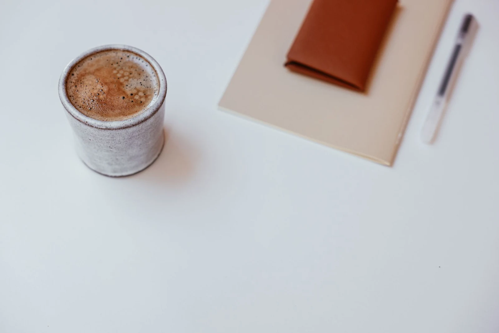 Xícara de café ao lado de dois cadernos fechados e uma caneta sobre uma mesa clara