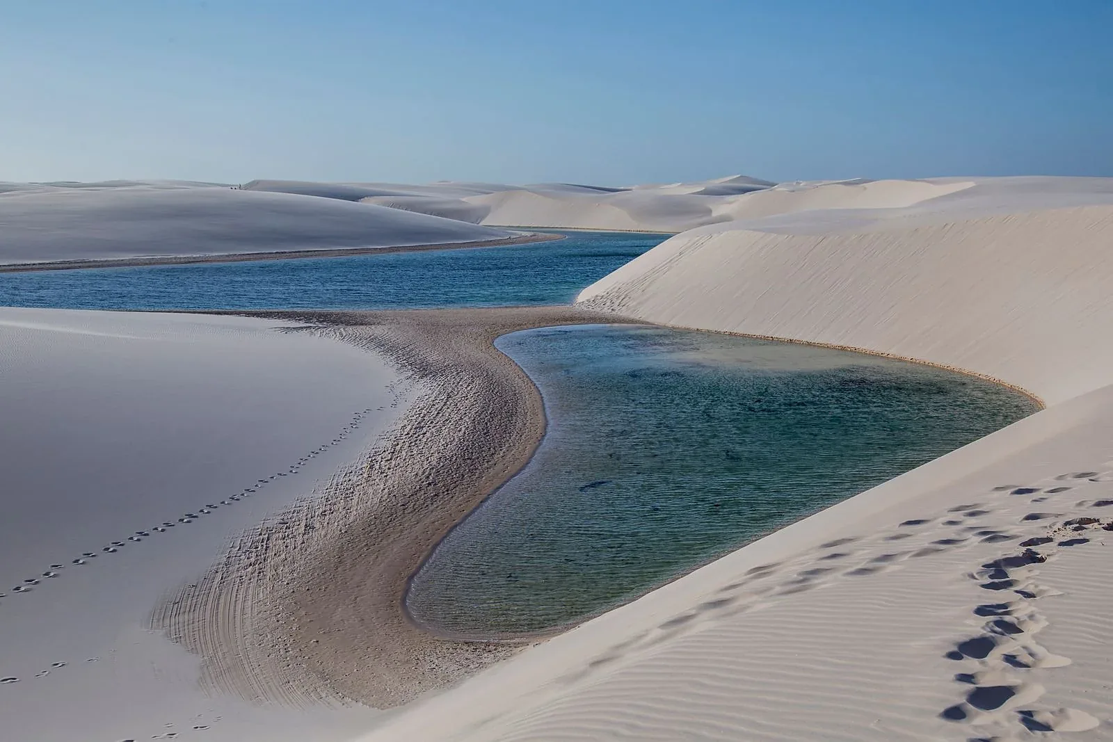 Dunas de areia branca com lagoas de água azul-esverdeada entre elas, pegadas na areia em primeiro plano e céu azul limpo