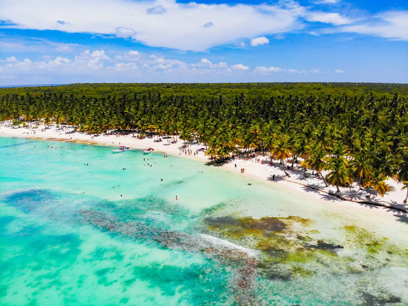 Vista aérea de faixa de areia branca com coqueiros e banhistas em água turquesa rasa, com manchas de recife visíveis no fundo do mar