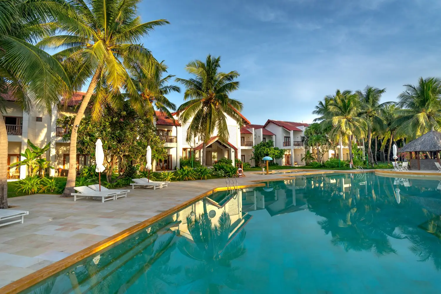 Piscina de resort ao lado de espreguiçadeiras brancas, guarda-sóis fechados e coqueiros altos em frente a prédios baixos de telhado vermelho