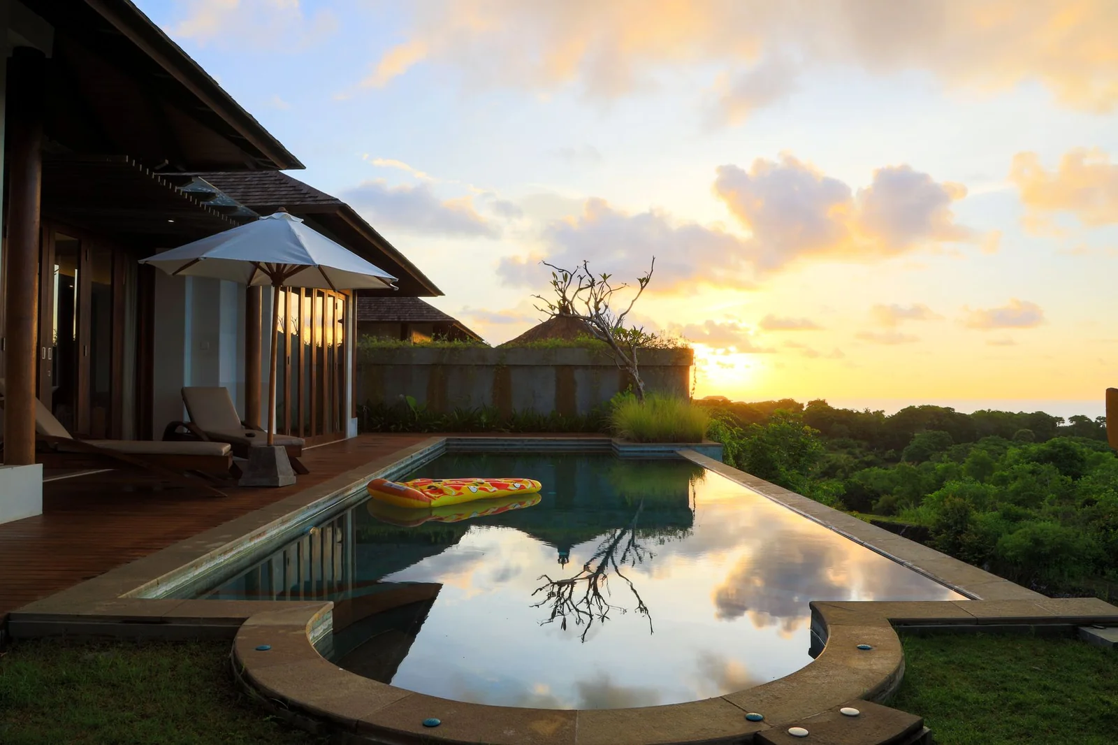 Piscina comprida de uma casa tropical ao entardecer, com deck de madeira, espreguiçadeira, guarda-sol branco e céu alaranjado