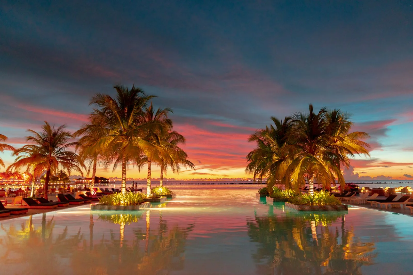 Piscina de resort ao entardecer com a água espelhando o céu rosa e alaranjado, dois grupos de coqueiros iluminados por luzes baixas nas laterais, espreguiçadeiras nas bordas e o mar calmo ao fundo