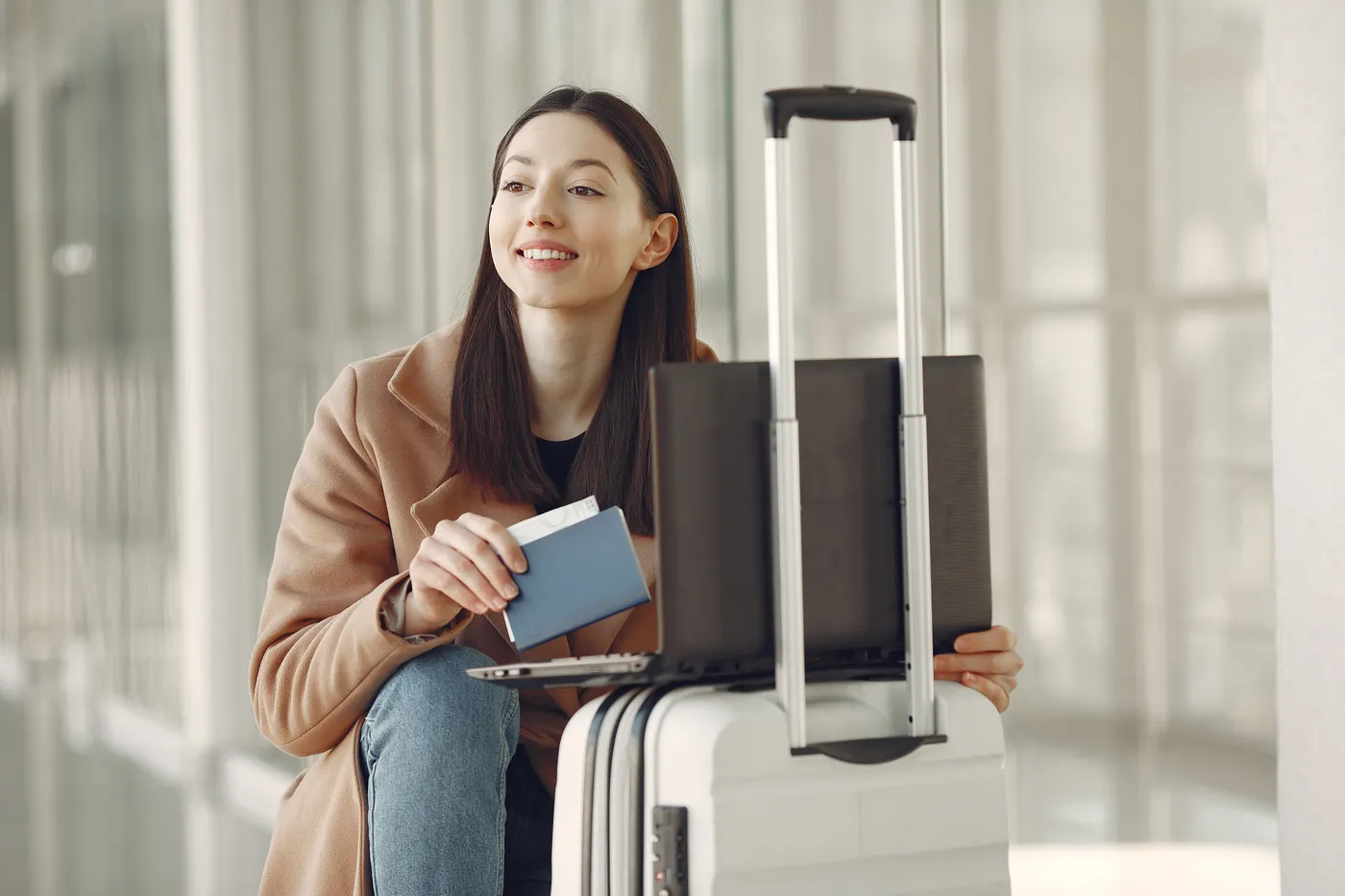 Mulher sorridente sentada em um aeroporto, com passaporte na mão e um notebook apoiado em uma mala branca
