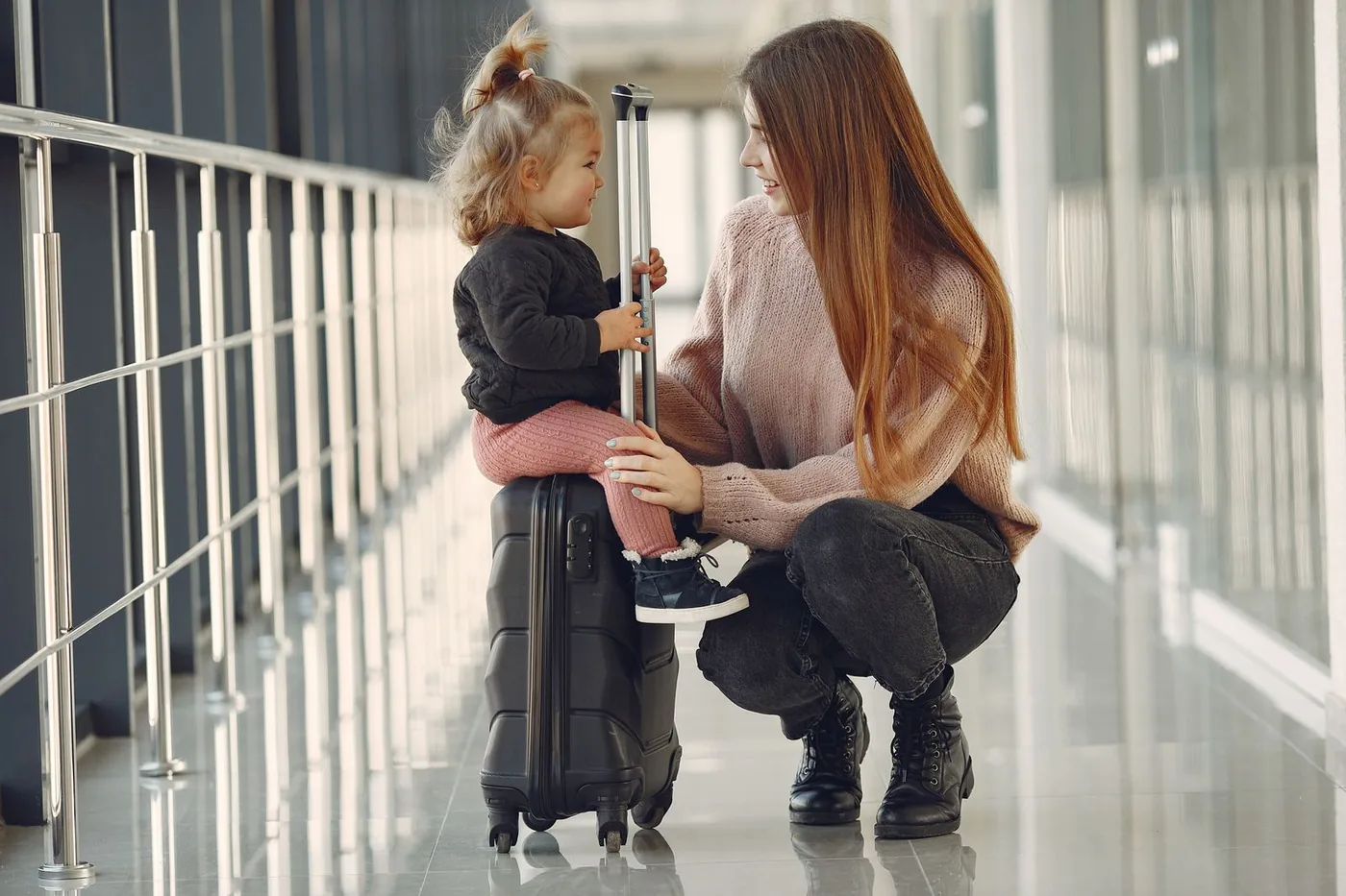 Mulher agachada ao lado de uma menina sentada sobre uma mala preta, em um corredor de aeroporto