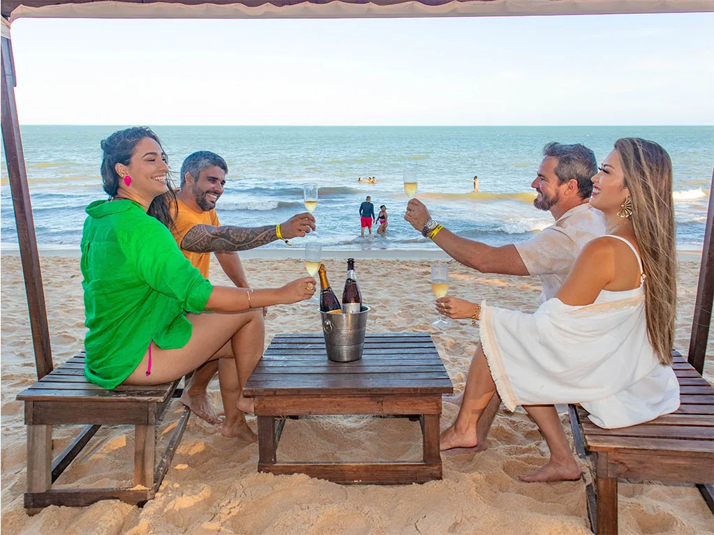 Dois casais brindam com espumante numa tenda na areia, de frente para o mar