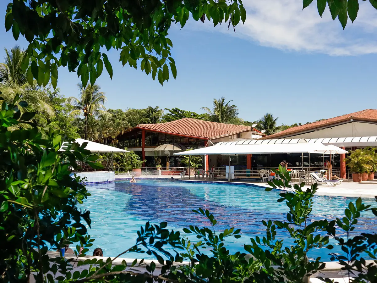 Piscina do Resort Arcobaleno entre árvores e coqueiros, com prédios de telhado vermelho ao fundo