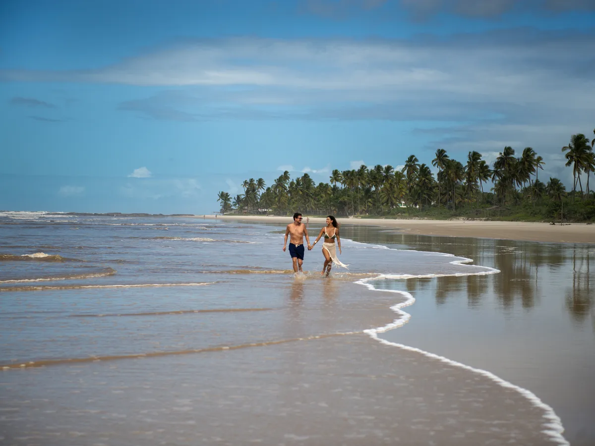 Casal caminhando de mãos dadas na beira do mar da praia de Canabrava, com coqueiros ao fundo
