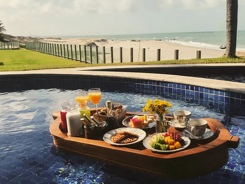 Café da manhã numa barca de madeira flutuando na piscina, com sucos, frutas e flores, e a praia ao fundo