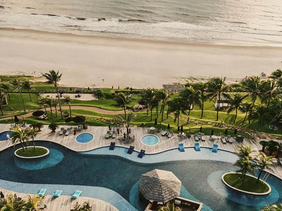 Vista aérea do Carmel Charme Resort, com a piscina e o bar molhado, o gramado com coqueiros e a praia larga em frente