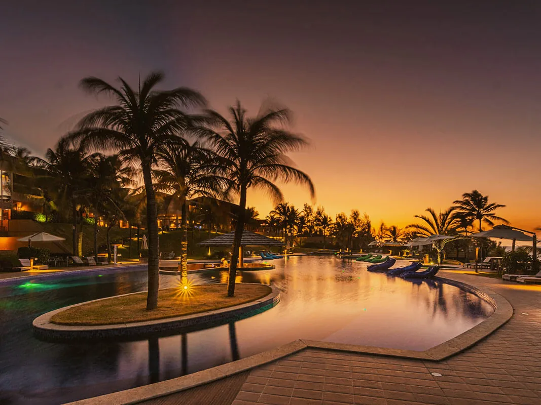 Piscina do Carmel Charme Resort ao entardecer, com coqueiros numa ilha e um quiosque de palha sobre a água