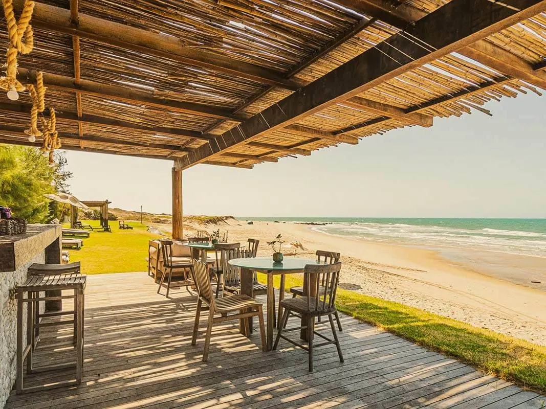 Deque coberto do bar da praia do Carmel Charme Resort, com mesas de madeira de frente para a areia e o mar