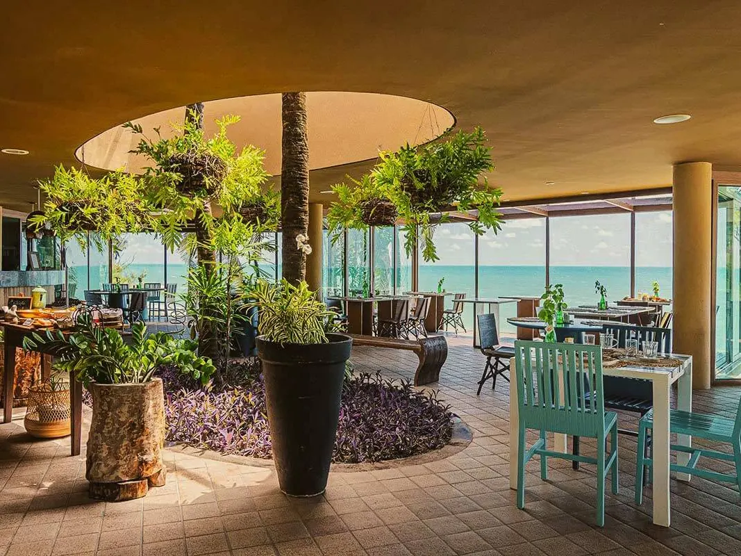 Salão do restaurante do Carmel Charme Resort, com mesas, plantas penduradas e janelas de vidro de frente para o mar
