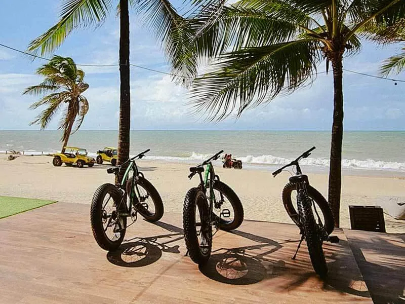 Três bicicletas de praia no deque do Carmel Cumbuco, com coqueiros, buggies na areia e o mar ao fundo