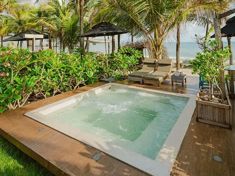 Jacuzzi num deque de madeira no jardim do Carmel Cumbuco, com espreguiçadeiras, coqueiros e o mar ao fundo