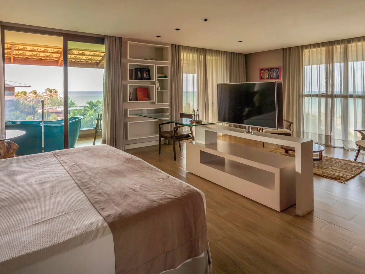 Suíte do Carmel Cumbuco com cama de casal, sala com TV e varanda de frente para o coqueiral e o mar