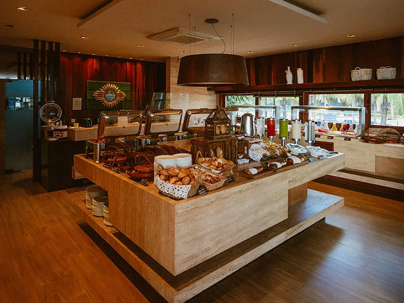 Bufê do café da manhã do Carmel Cumbuco, com pães, frutas e sucos num balcão de madeira