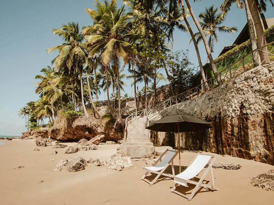 Praia da Taíba ao pé do rochedo do resort, com duas cadeiras de praia sob um guarda-sol e a escada de pedra que desce até a areia