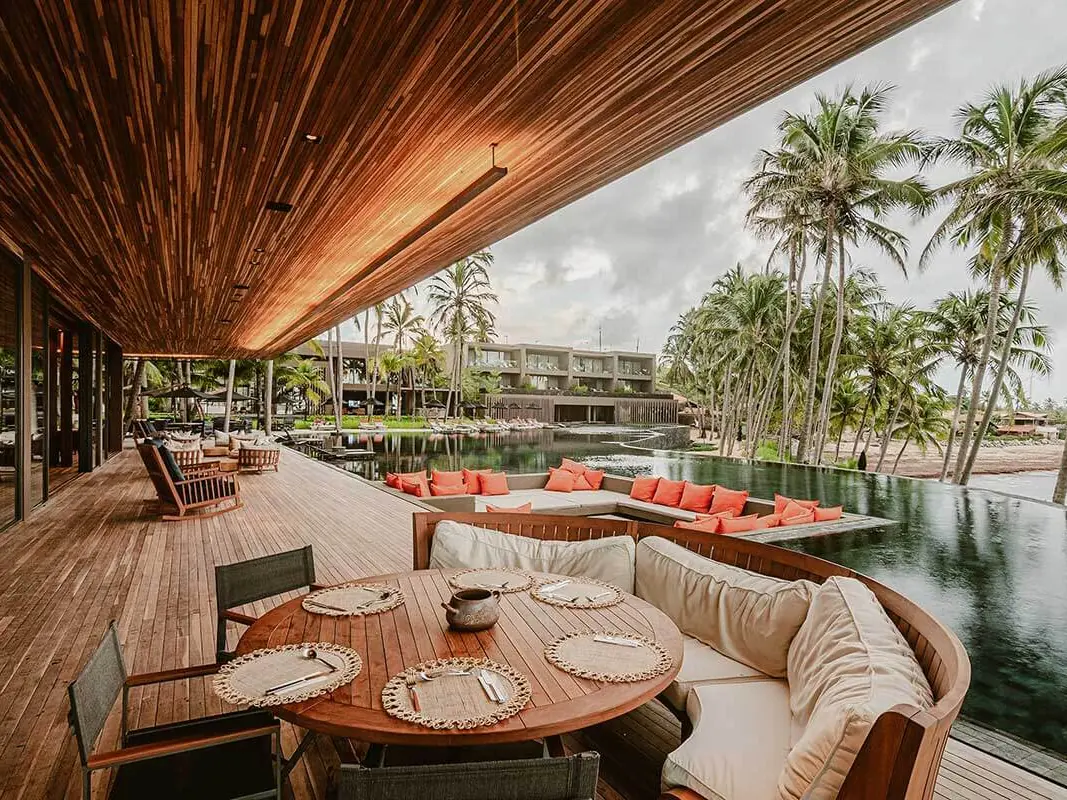 Varanda do Restaurante Cipó, com mesa redonda posta ao lado da piscina de borda infinita e coqueiros ao fundo