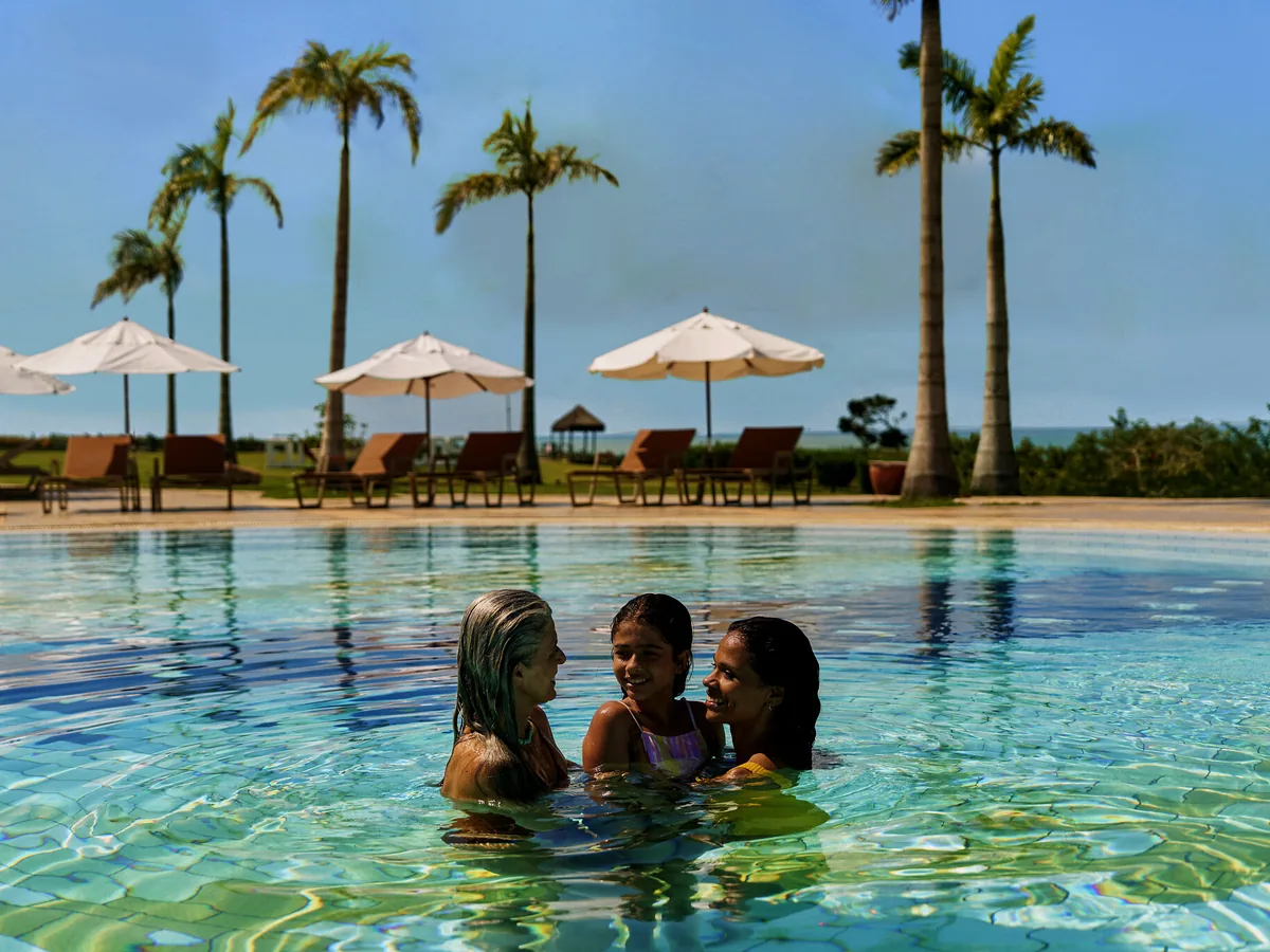 Família dentro da piscina do Club Med Trancoso, com palmeiras, guarda-sóis e espreguiçadeiras ao fundo