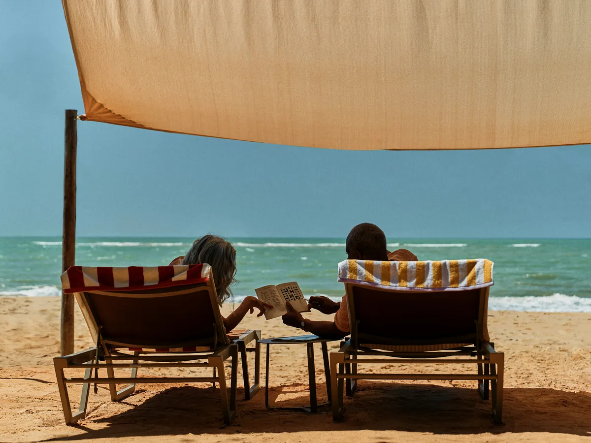 Casal em espreguiçadeiras sob um toldo na areia, de frente para o mar