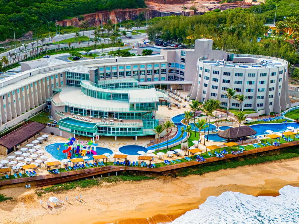 Vista aérea do Costeira Palace, com a Via Costeira atrás dos prédios, as piscinas, o parque aquático infantil e a praia em frente
