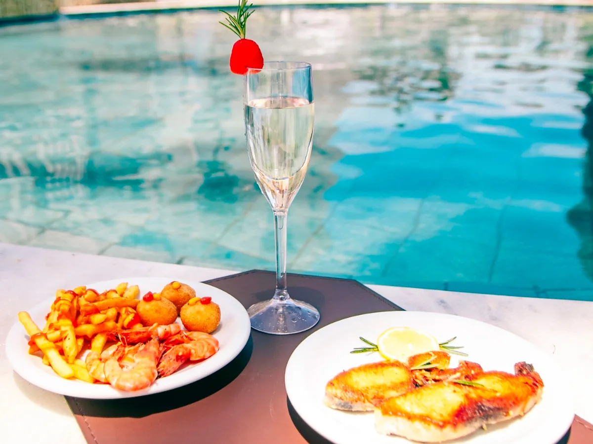 Petiscos, peixe grelhado e taça de espumante na borda da piscina do Costeira Palace