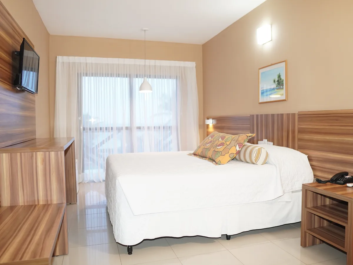 Quarto do El Aram Imirá com cama de casal, TV na parede e porta de vidro com cortina clara