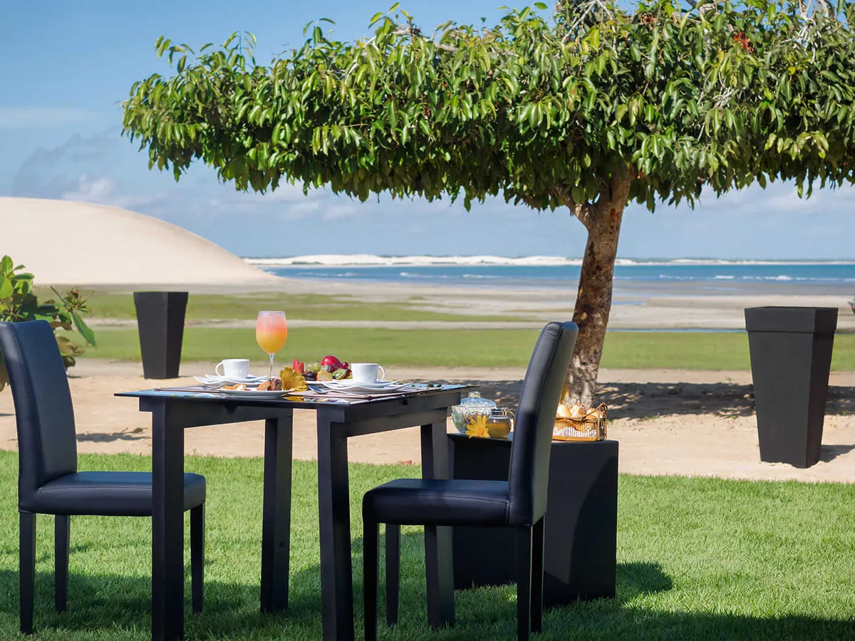 Mesa de café da manhã no gramado do Essenza Hotel, à sombra de uma árvore, com a praia e a duna ao fundo