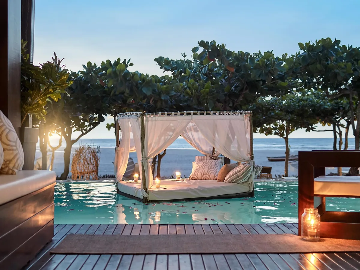 Cama de dossel com velas à beira da piscina do Essenza Hotel ao entardecer, com árvores e o mar ao fundo