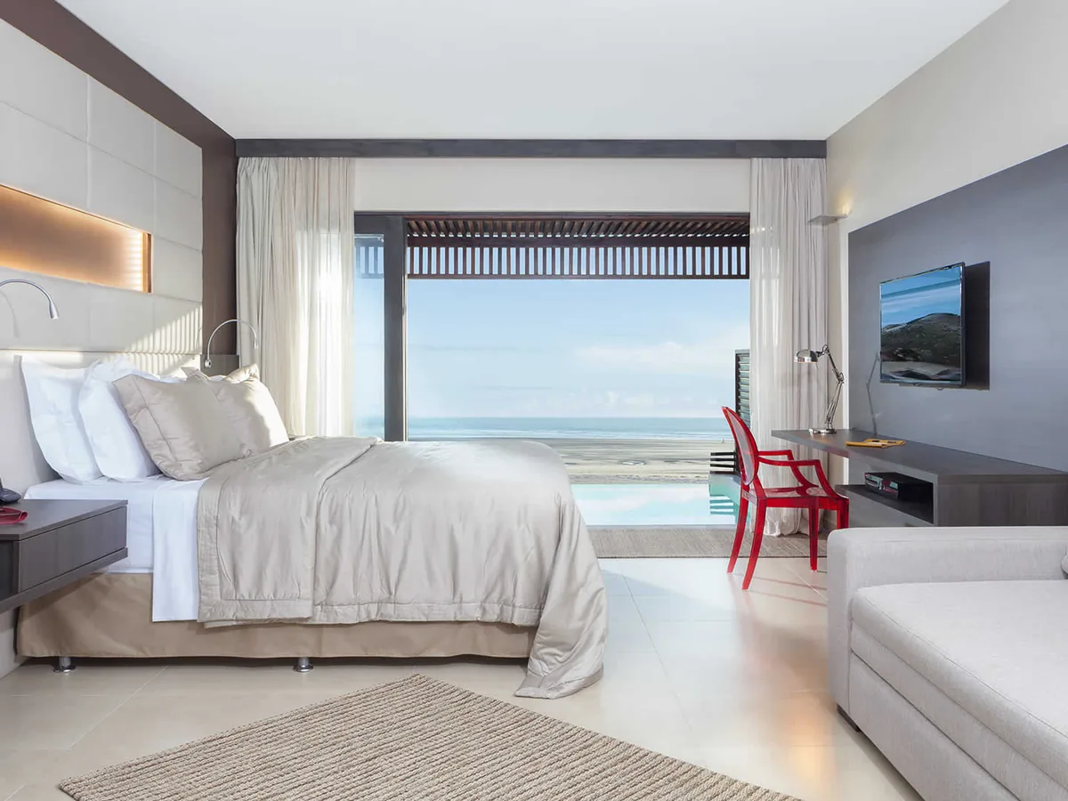 Apartamento do Essenza Hotel com cama king, sofá e porta de vidro aberta para a piscina e o mar