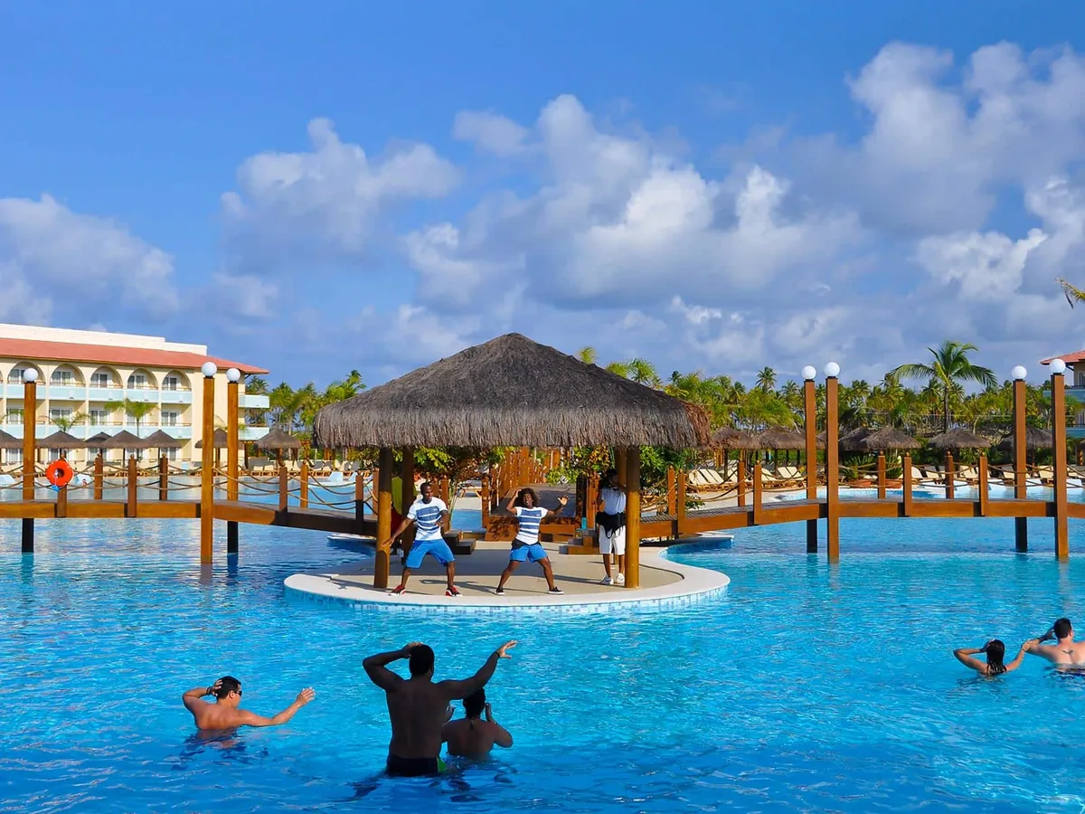 Hóspedes fazendo aula de ginástica dentro da piscina principal do Grand Palladium Imbassaí, com monitores num quiosque de palha