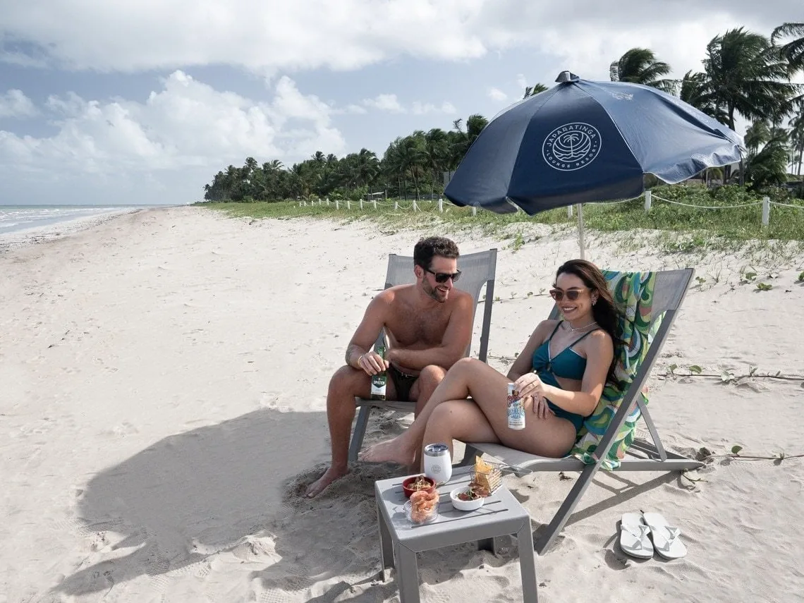 Casal em cadeiras de praia sob o guarda-sol do Japaratinga Lounge Resort, na areia da praia de Japaratinga