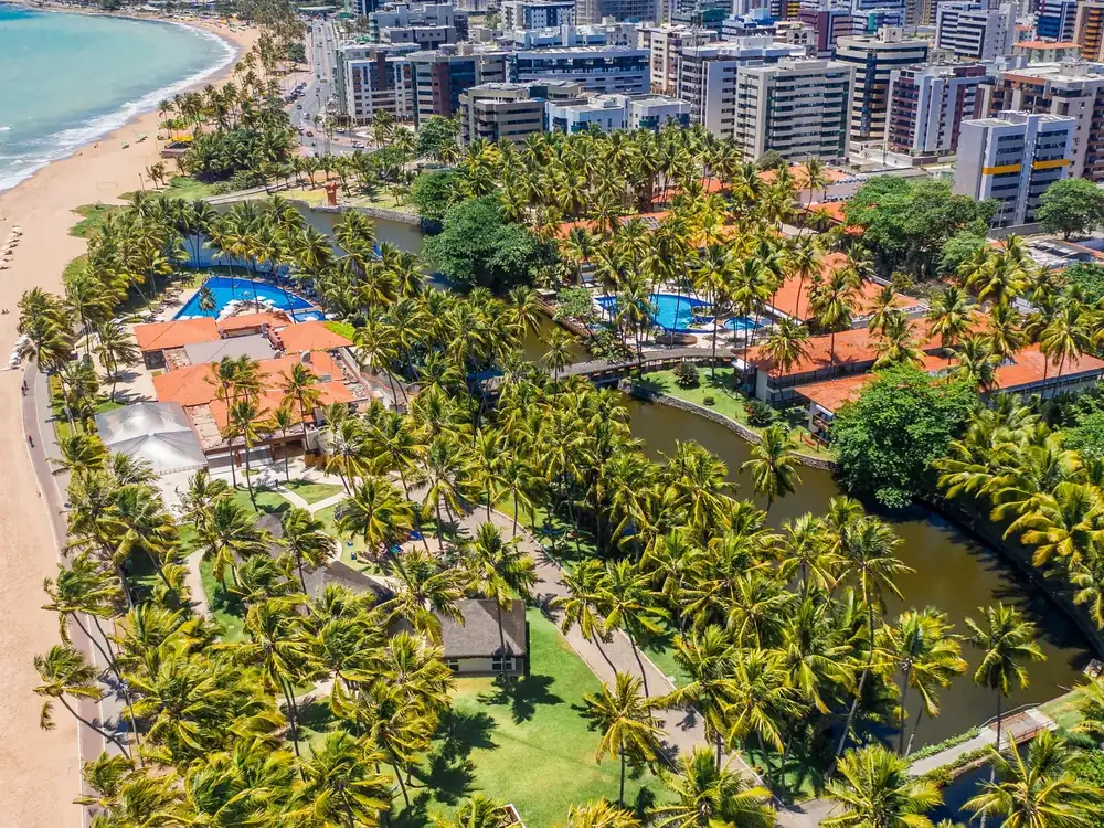 Vista aérea do Jatiúca Resort na orla de Maceió, com a praia, o coqueiral, as piscinas, a lagoa e os prédios da cidade ao lado