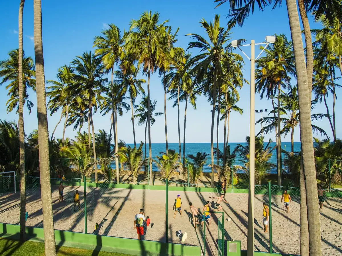 Hóspedes jogando futebol na quadra de areia do Jatiúca Resort, entre coqueiros, com o mar ao fundo
