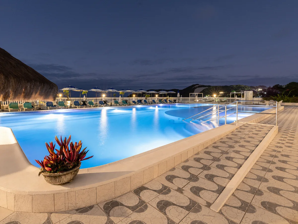 Piscina no terraço do SERHS Beach Hotel ao anoitecer, iluminada, com guarda-sóis e o Morro do Careca ao fundo