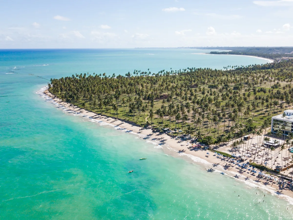 Vista aérea do litoral de Ipioca, com o mar verde, a praia, o coqueiral e o prédio do Maceió Mar Resort à direita