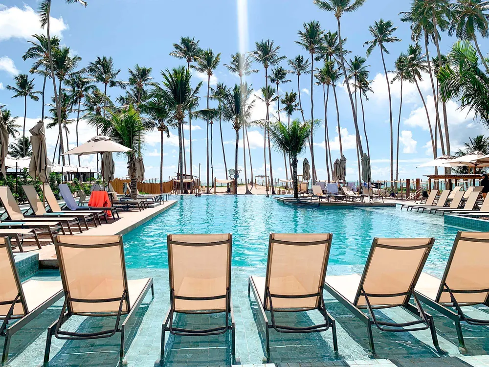 Piscina do Maceió Mar Resort com espreguiçadeiras dentro d'água e coqueiros ao fundo