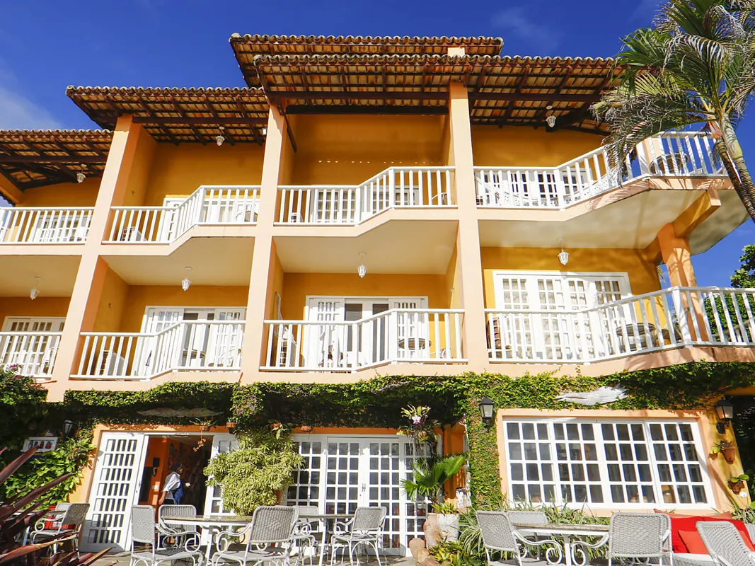 Fachada neocolonial do Manary Praia Hotel, com varandas brancas, telhado de telhas e mesas no jardim
