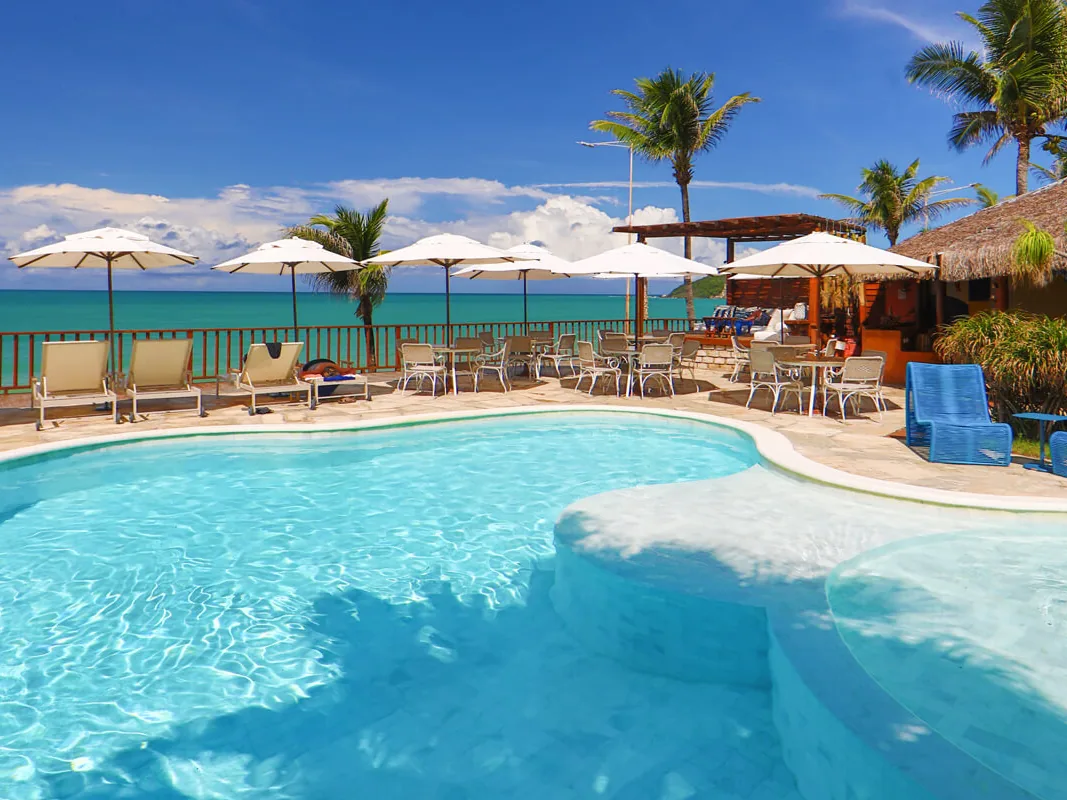 Piscina do Manary Praia Hotel com espreguiçadeiras e guarda-sóis brancos, com o mar verde ao fundo