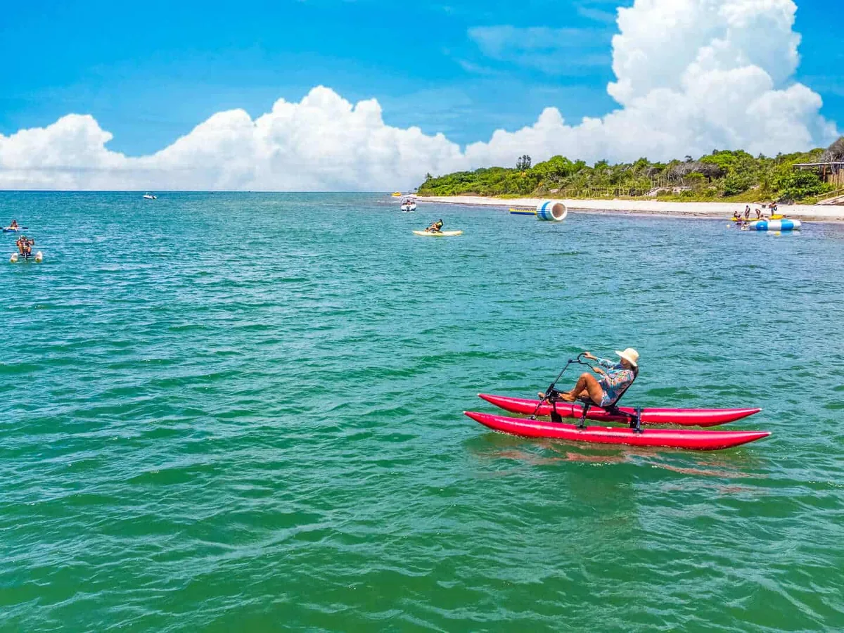 Hóspede numa bicicleta aquática no mar da Praia do Mutá, com caiaques e boias do Beach Club Paradiso ao fundo