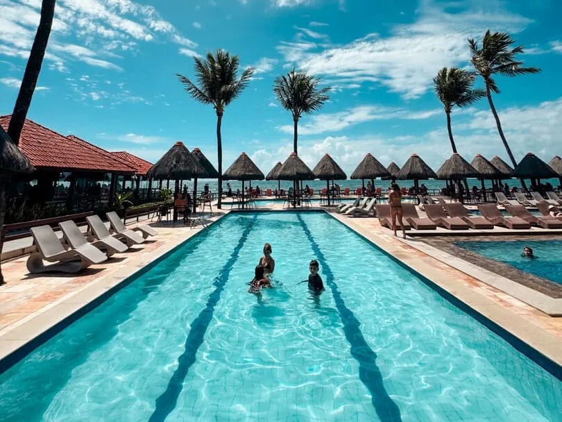 Família nadando na piscina de raias do Ocaporã Hotel, com quiosques de palha e coqueiros ao fundo