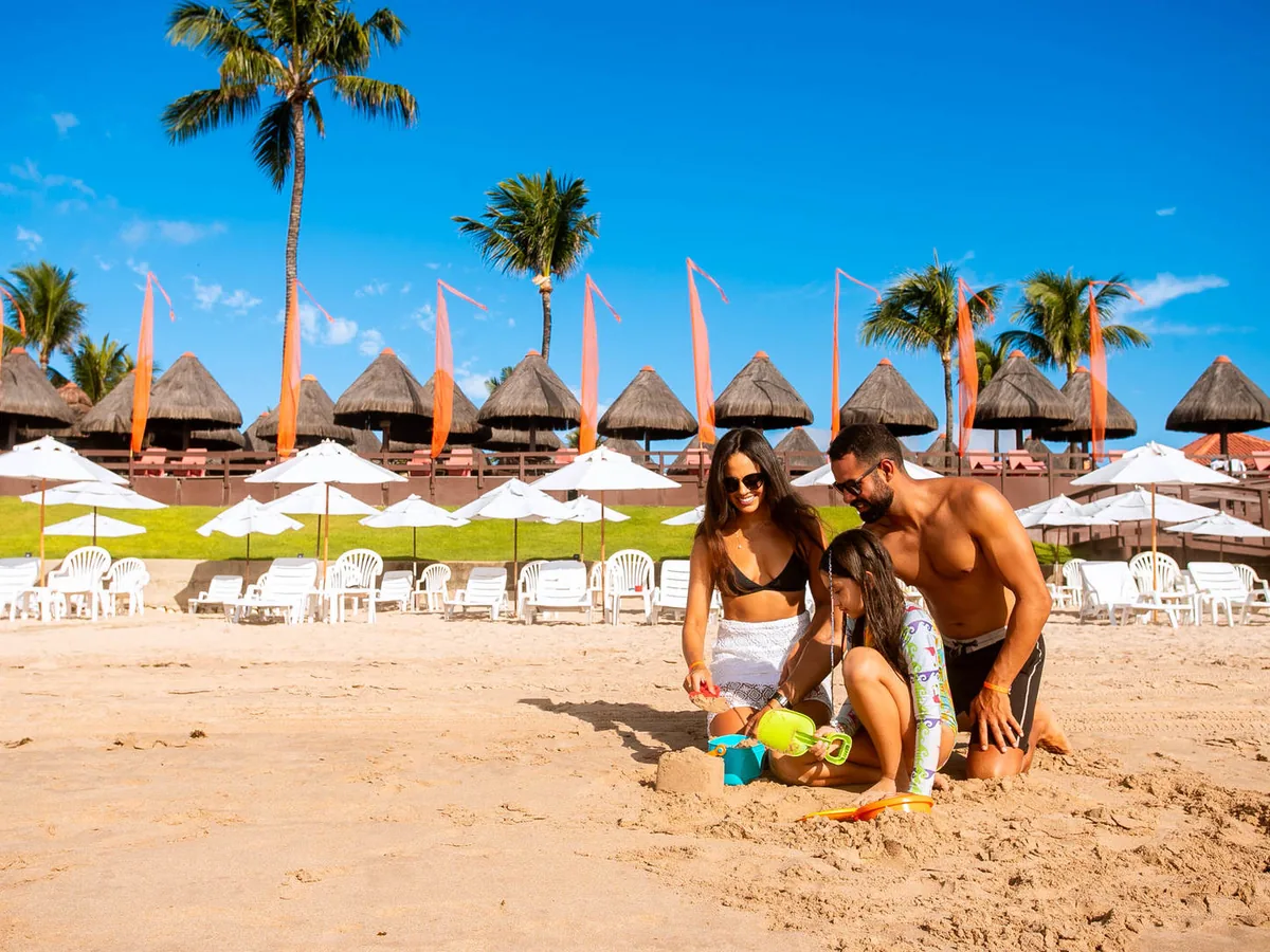 Família fazendo castelo de areia na Praia do Cupe, com os quiosques de palha do Ocaporã Hotel ao fundo