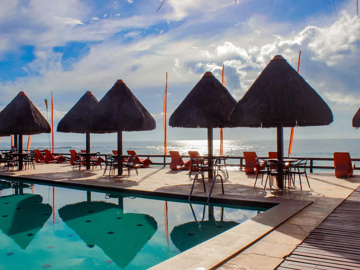 Piscina do Ocaporã Hotel com quiosques de palha na borda e o mar da Praia do Cupe logo atrás