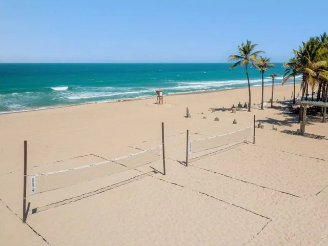 Praia em frente ao Oceani Beach Park, com redes de vôlei na areia, coqueiros e o mar