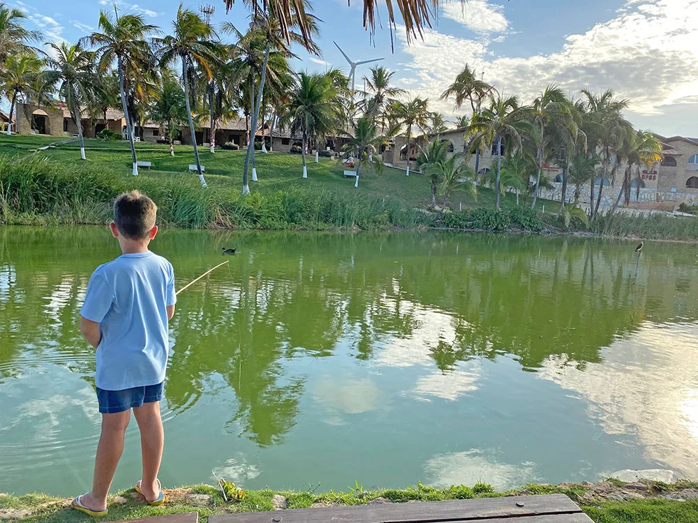 Menino pescando de vara no lago do Hotel Parque das Fontes, com coqueiros e os prédios do hotel na outra margem