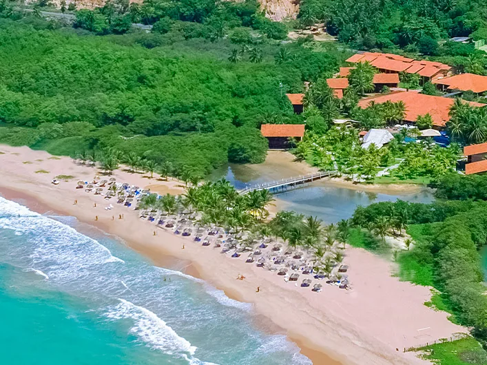 Vista aérea do Pratagy Beach Resort, com a praia, os guarda-sóis, a passarela sobre o rio e os telhados entre os coqueiros
