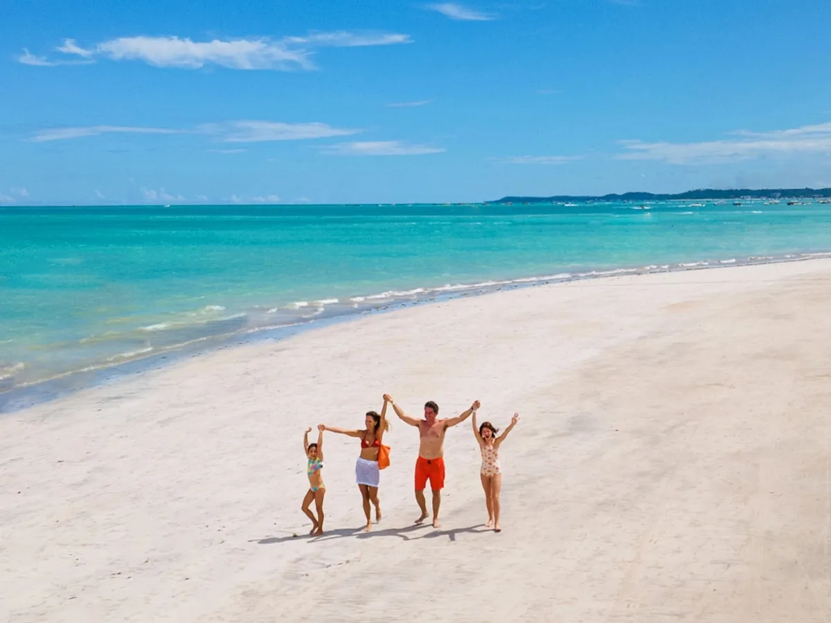 Família de mãos dadas na areia branca da praia de Maragogi, com o mar azul ao fundo